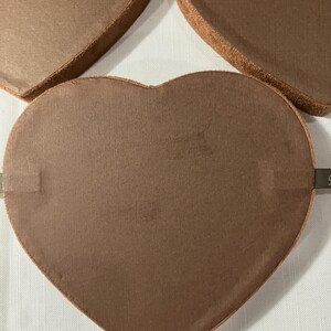 May include: Close-up of several heart-shaped boxes in a warm brown color. The boxes have a textured surface and a darker brown trim. The boxes are arranged on a white surface.