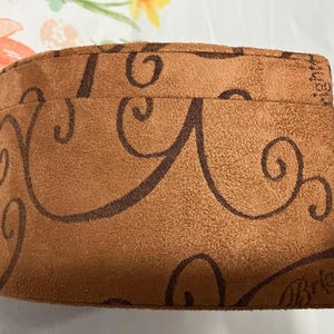 May include: A brown pouch with a suede-like texture, adorned with dark brown swirling patterns. The pouch has a drawstring closure and the word "Brighton" is visible on the side. The pouch is on a floral patterned surface.