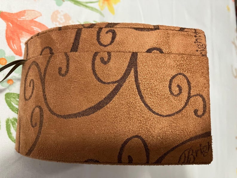 May include: A brown pouch with a suede-like texture, adorned with dark brown swirling patterns. The pouch has a drawstring closure and the word "Brighton" is visible on the side. The pouch is on a floral patterned surface.