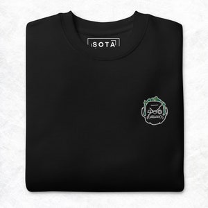 May include: Black t-shirt with a white embroidered design of a cartoon face with green hair. The text "ISOTA" is embroidered on the shirt.