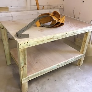May include: A wooden workbench with a single shelf. The workbench is made of unfinished wood and has a light brown color. There are two yellow straps and a metal piece on the top shelf.