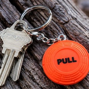 May include: A silver-colored metal key ring holding two keys, one of which is marked with the word SCHLAGE, and a bright orange circular clay target replica keychain with the word PULL printed in black letters on the front surface.