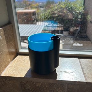 Könnte beinhalten: Ein zweifarbiger Becher mit eingebautem Halter. Der obere Teil des Bechers ist hellblau, während der untere Teil und der Halter schwarz sind. Der Becher steht auf einer beigefarbenen Fliesenoberfläche.