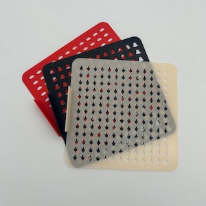 May include: Three square stencils in red, black, and off-white. The red stencil has evenly spaced holes. The black stencil has leaf-shaped cutouts. The off-white stencil has leaf-shaped cutouts in black and red.