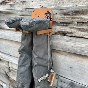 May include: A wooden boot rack with a decorative tree and bird design, holding a pair of gray waders. The rack is mounted on a weathered wooden wall, with a chain and accessories hanging from the side.