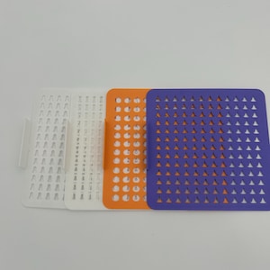May include: Three colorful plastic stencils. The first is white, the second is orange, and the third is purple. Each stencil has a different pattern of cut-out shapes. The stencils are rectangular with rounded corners.