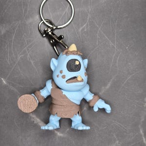 May include: A blue cyclops keychain with a single eye, a brown loincloth, and a circular object in its hand. The keychain has a silver ring and clasp. The cyclops has a light blue body and a brown textured design.