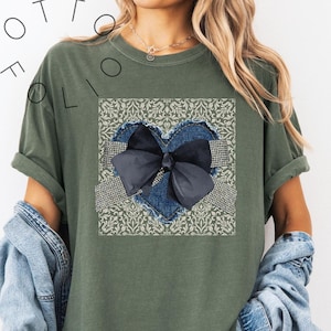 Puede incluir: Camiseta verde oliva con un diseño en forma de corazón. El corazón tiene un contorno de mezclilla y un lazo azul oscuro, sobre un fondo estampado. El texto "OTTO FOLIO" está impreso en la camiseta.