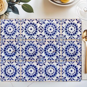 May include: A rectangular tile trivet composed of twelve square tiles. Each tile features a blue and white floral design with a central medallion and surrounding scrollwork. The trivet is placed on a white surface, with gold cutlery and a flower visible.