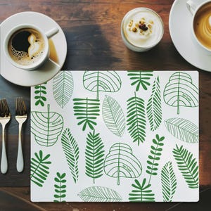 Puede incluir: Mantel individual blanco con un patrón de hojas verdes. El diseño presenta varias formas de hojas y el texto "FUCK TRUMP". El mantel individual está sobre una mesa de madera, con tazas de café y un cuenco pequeño de crema.