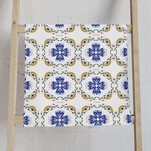 May include: White towel with a repeating floral pattern in blue, yellow, and green. The design features stylized blue flowers, yellow petal-like shapes, and green leaf accents. The towel is draped over a wooden rack.