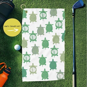 May include: White golf towel with a repeating pattern of green turtles. The towel has a loop for hanging and is displayed on a green surface with golf equipment and a yellow circle with the text "ANTI-TRUMP GIFT".