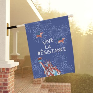 Peut inclure: Un drapeau bleu avec le texte "VIVE LA R&Eacute;SISTANCE" et des illustrations de feux d'artifice, de la Statue de la Libert&eacute; et de drapeaux am&eacute;ricains. Le drapeau est accroch&eacute; &agrave; l'ext&eacute;rieur sur un poteau blanc.