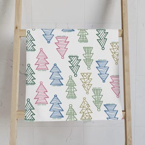 Puede incluir: Toalla blanca con un patrón repetido de árboles de Navidad estilizados en verde, azul, rosa y dorado. La frase "FUCK TRUMP" está impresa debajo de cada árbol. La toalla está colgada en una escalera de madera.