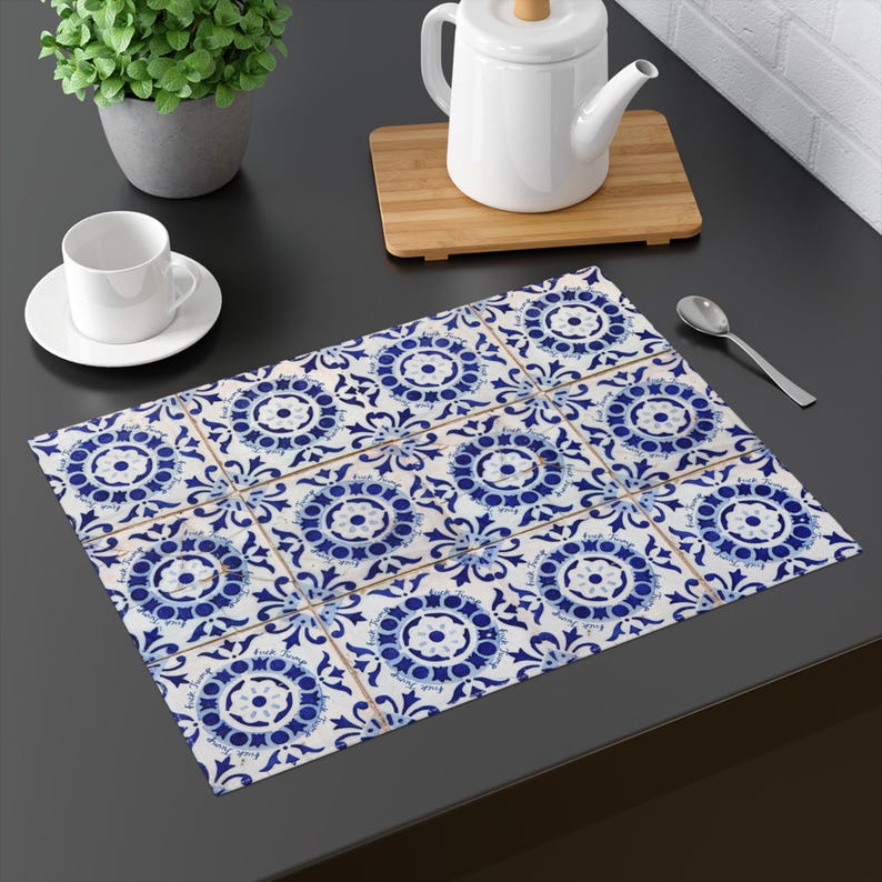 Pu&ograve; includere: Un sottopiatto rettangolare con un motivo ripetuto di piastrelle floreali blu e bianche. Il sottopiatto &egrave; su una superficie scura, con una tazza e un piattino bianchi, una teiera e una piccola pianta.