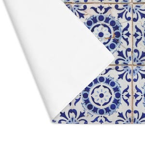 Pu&ograve; includere: Un sottopiatto bianco con un angolo ripiegato, che rivela una sezione di piastrelle a motivi blu e bianchi. Le piastrelle hanno un design floreale e le parole "Fuck Trump" sono stampate su di esse.