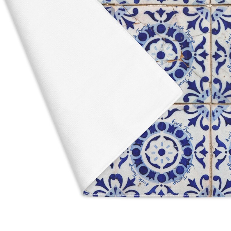 Pu&ograve; includere: Un sottopiatto bianco con un angolo ripiegato, che rivela una sezione di piastrelle a motivi blu e bianchi. Le piastrelle hanno un design floreale e le parole "Fuck Trump" sono stampate su di esse.