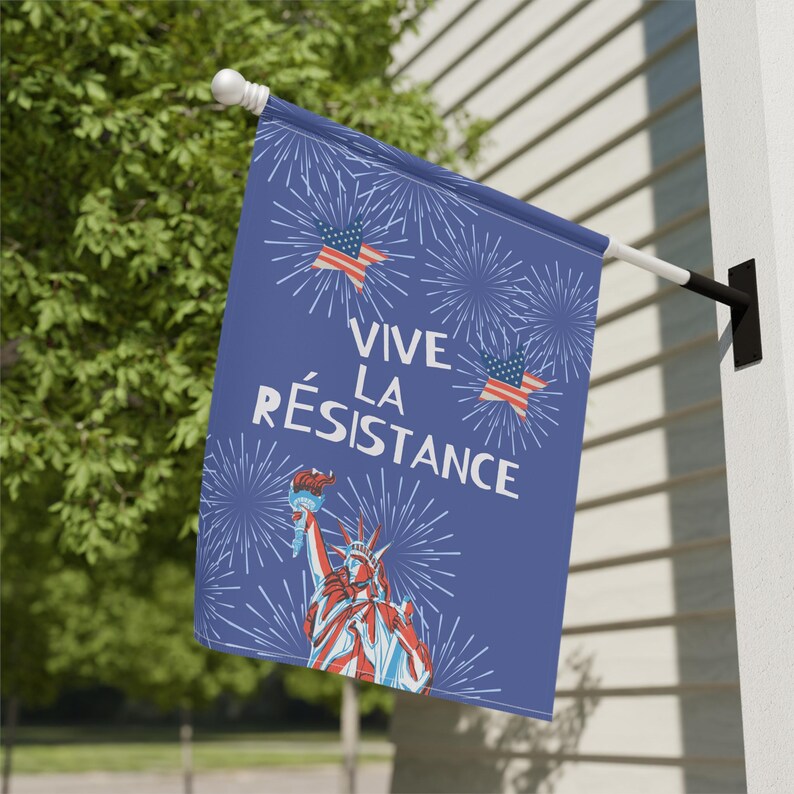 Peut inclure: Un drapeau bleu avec le texte "VIVE LA R&Eacute;SISTANCE" et des illustrations de la Statue de la Libert&eacute;, des drapeaux am&eacute;ricains et des feux d'artifice. Le drapeau est accroch&eacute; &agrave; l'ext&eacute;rieur sur un mur blanc.