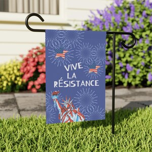 Peut inclure: Un drapeau de jardin bleu avec le texte "VIVE LA R&Eacute;SISTANCE" et des illustrations de la Statue de la Libert&eacute;, des drapeaux am&eacute;ricains et des feux d'artifice. Le drapeau est affich&eacute; sur un support en m&eacute;tal noir dans un jardin.