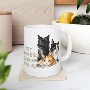 May include: White ceramic mug featuring a design of three cats: a black cat, a black and white cat, and an orange and white cat. The mug has the text "YET ANOTHER REASON LOVE CATS".