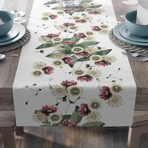 May include: White table runner with a floral design featuring burgundy and cream-colored flowers, green leaves, and small bee illustrations. The runner is placed on a wooden table, with place settings visible on either side.