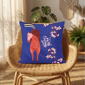 May include: Blue decorative pillow featuring a stylized red horse with pink accents and the year 2026. The pillow also includes floral branches and Chinese characters. The pillow is placed on a wicker chair.