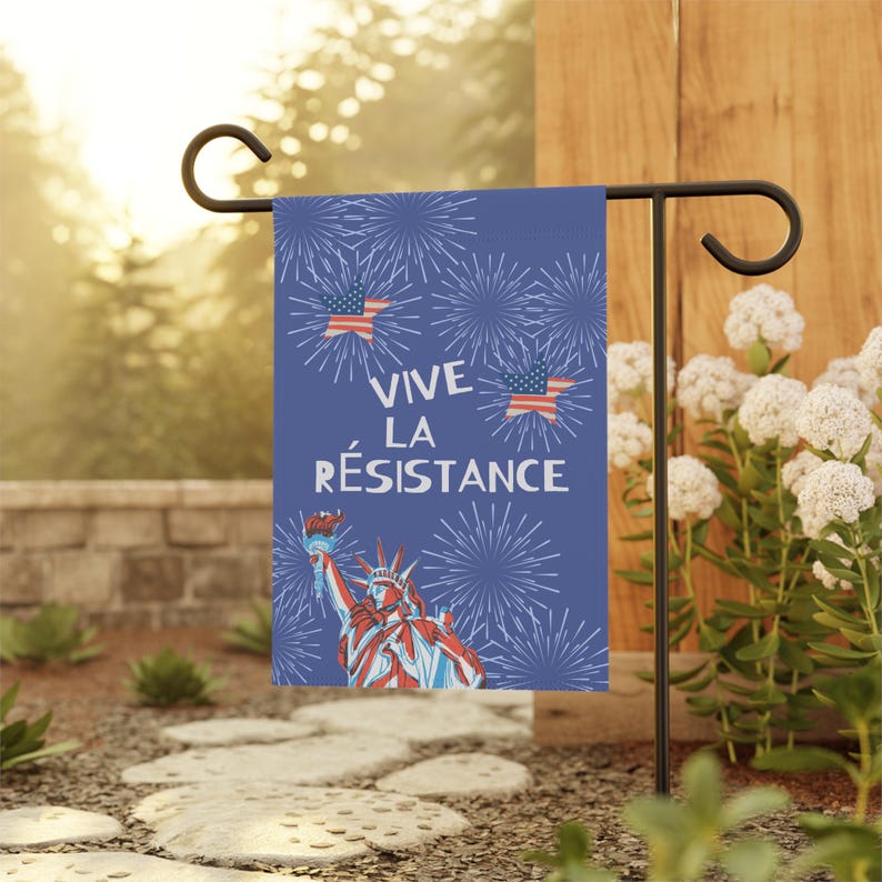 A blue garden flag with the text "Vive la Résistance" (Long Live the Resistance in French) and an illustration of the Statue of Liberty. The flag also features American flags and fireworks and is meant to be a patriotic Anti Trump message. The flag is displayed on a black metal stand.