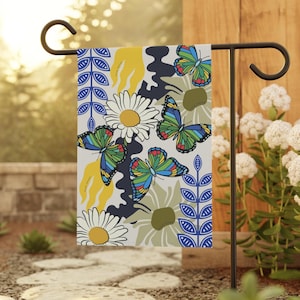 Può includere: Una bandiera da giardino decorativa con un motivo a farfalle e margherite. La bandiera ha uno sfondo grigio chiaro con accenti blu, gialli e verdi. È esposta su un supporto in metallo nero.