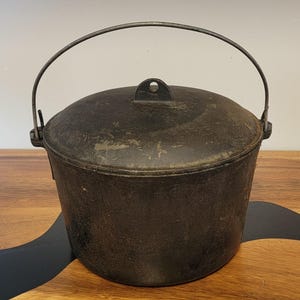 May include: A black cast iron Dutch oven with a lid and a metal handle. The pot has a round shape and is designed for cooking. The handle arches over the top of the pot. The pot is sitting on a wooden surface.