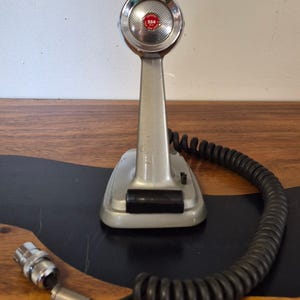 Puede incluir: Un micrófono de escritorio plateado con una cabeza circular que presenta un emblema central rojo con la etiqueta 254. El micrófono tiene un soporte cónico, un interruptor rectangular negro en la base y un largo cable negro en espiral.