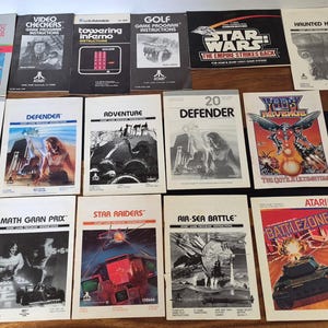 Può includere: Una collezione di diciassette manuali di istruzioni per videogiochi Atari vintage disposti in una griglia. Gli opuscoli presentano vari titoli tra cui E.T., Star Wars, Defender e Missile Command, con un mix di copertine in bianco e nero e a colori.
