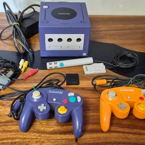 Peut inclure: Une console Nintendo GameCube violette avec une façade argentée, deux manettes filaires violette et orange, un bloc d'alimentation noir, un câble AV, un microphone blanc, une carte mémoire et un adaptateur gris. Le logo GameCube est visible.
