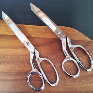 May include: Two pairs of silver-colored metal sewing scissors with polished blades and ergonomic handles. Each blade features a visible engraved logo and text. The scissors are positioned side-by-side, showcasing their sharp, pointed tips and sturdy construction for precise cutting tasks.