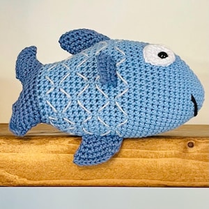 Könnte beinhalten: Ein gehäkeltes blaues Fischspielzeug mit weißen Nähten und einem schwarzen Auge. Der Fisch liegt auf einer Holzoberfläche.