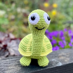 May include: A crocheted green turtle toy with white eyes. The turtle has a striped shell and is sitting on a wooden surface.