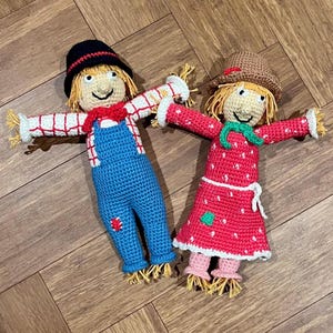 May include: Two handmade crochet scarecrows. One wears blue overalls, a red and white plaid shirt, and a black hat. The other wears a red dress with white polka dots, a brown hat, and pink leggings. Both have straw arms and legs.