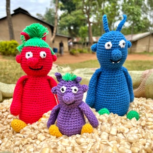 May include: Three crocheted stuffed toys with googly eyes. The toys are a red monster, a blue monster, and a purple monster. The toys are sitting on a bed of corn kernels.
