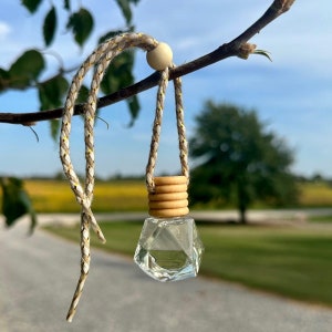 May include: A clear glass car air freshener with a geometric shape and a wooden cap. The freshener is hanging from a beige and gold rope.
