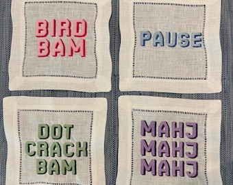 May include: Four square white linen coasters with embroidered text in various colors. The coasters feature the words "BIRD BAM", "PAUSE", "DOT CRACH BAM", and "MAHJ MAHJ MAHJ". Each coaster has a black stitched border.