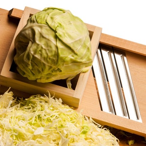 May include: A wooden cabbage shredder with a green cabbage in the holder and shredded cabbage in the foreground. The shredder has a metal blade and the text "CabbageShredder.com" is printed on the side.