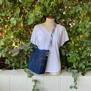 May include: A denim tote bag with a floral patterned strap. The bag is hanging on a white mannequin wearing a white t-shirt. The mannequin is standing in front of a green leafy bush.