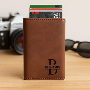 May include: A brown leather wallet with a personalized design. The wallet has a monogram with the letter "D" and the name "Benjamin" underneath. Credit cards are visible inside the wallet. The wallet is on a wooden surface.