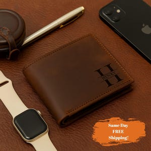 May include: A brown leather wallet with the name "Henry" and the initial "H" engraved on the front. A gold pen, a black smartphone, and a smartwatch with a cream-colored band are also visible.