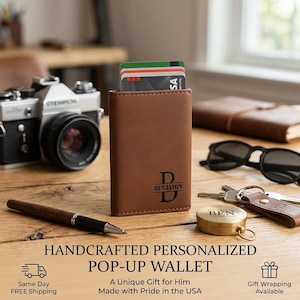May include: A brown leather pop-up wallet featuring a black engraved monogram with the letter D and the name Benjamin. The wallet is standing upright and contains several credit cards. It is positioned on a wooden surface with a pen and keys.