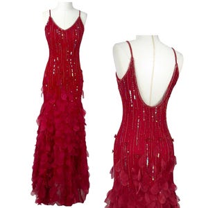 Könnte beinhalten: Ein bodenlanges rotes Kleid mit dünnen Spaghettiträgern, einem V-Ausschnitt und einem tiefen Rückenausschnitt. Das Oberteil ist mit vertikalen Perlenreihen und Pailletten verziert, während der Rock mit geschichteten, blütenblattartigen Applikationen versehen ist.