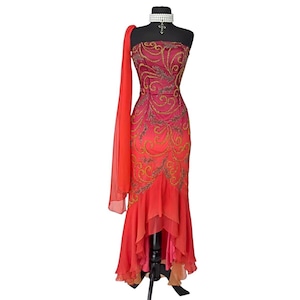 May include: A red and orange ombre strapless gown with gold embroidery and a high-low hemline. The gown is on a mannequin and has a sheer scarf draped over one shoulder.