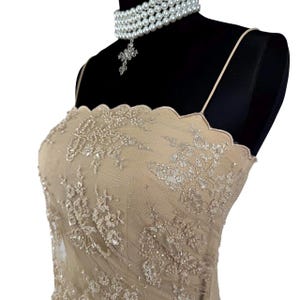 Puede incluir: Vestido de fiesta beige adornado, con escote festoneado y tirantes finos. El vestido est&aacute; adornado con intrincados bordados florales y lentejuelas brillantes. Un collar de perlas de varias hebras con un colgante de cruz se usa en un maniqu&iacute; negro.