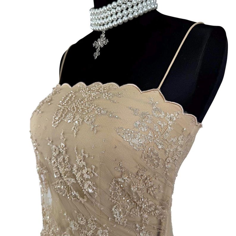 Puede incluir: Vestido de fiesta beige adornado, con escote festoneado y tirantes finos. El vestido est&aacute; adornado con intrincados bordados florales y lentejuelas brillantes. Un collar de perlas de varias hebras con un colgante de cruz se usa en un maniqu&iacute; negro.