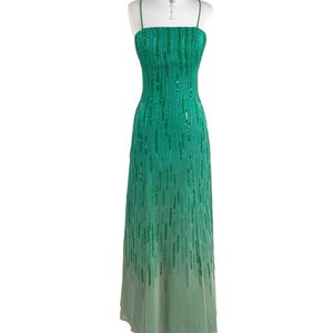 Könnte beinhalten: Ein bodenlanges smaragdgrünes Kleid mit dünnen Spaghettiträgern und geradem Ausschnitt. Das Kleid ist mit vertikalen Reihen aus schimmernden grünen Pailletten verziert, die am Saum in einen helleren, einfarbigen hellgrünen Stoff übergehen.