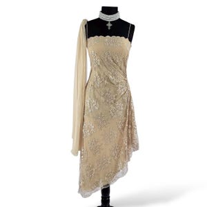 Puede incluir: Vestido de noche beige asim&eacute;trico con bordado floral y una manga de gasa fluida. El vestido se exhibe en un maniqu&iacute; negro y se combina con un collar de gargantilla de perlas.
